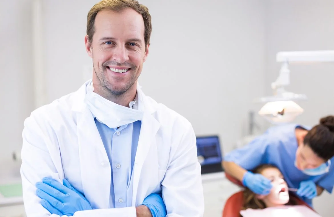 Klinikte kollarını kavuşturmuş bir şekilde duran gülümseyen diş hekimi